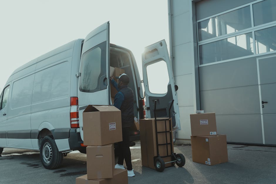 A man wearing a cap and a dark jacket is standing beside the open rear doors of a large white commercial van, which is parked outside a modern building with a metallic facade and large glass windows. The man appears to be handling cardboard boxes, some inside the van and others on the pavement. Several unpacked cardboard boxes, some sealed with tape and others open, are placed on the ground near the van. An orange hand truck or dolly with a black handle is positioned next to the man, ready for moving items. The scene is outdoors, with bright natural lighting indicating daylight, suggesting a home relocation or furniture transport process involving careful packing and loading of household belongings. The environment appears organized, with the van loading process in progress, reflecting professional removals services such as those offered by Man and Van Chelsea, supporting efficient house removals in the SW3 Sloane Square area.