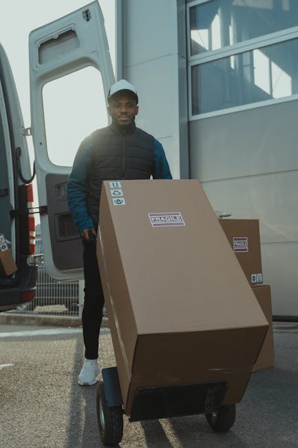 A man wearing a cap and a dark jacket is standing beside the open rear doors of a large white commercial van, which is parked outside a modern building with a metallic facade and large glass windows. The man appears to be handling cardboard boxes, some inside the van and others on the pavement. Several unpacked cardboard boxes, some sealed with tape and others open, are placed on the ground near the van. An orange hand truck or dolly with a black handle is positioned next to the man, ready for moving items. The scene is outdoors, with bright natural lighting indicating daylight, suggesting a home relocation or furniture transport process involving careful packing and loading of household belongings. The environment appears organized, with the van loading process in progress, reflecting professional removals services such as those offered by Man and Van Chelsea, supporting efficient house removals in the SW3 Sloane Square area.