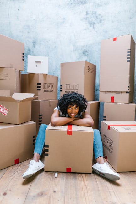 A young woman with curly black hair, dressed in a grey top, blue jeans, and white sneakers, is sitting on the wooden floor inside a room surrounded by numerous cardboard boxes of varying sizes, some with red and black tape seals. She is smiling and resting her arms on a large cardboard box in front of her, with her legs spread out and crossed at the ankles. The boxes are arranged around her, some stacked against a light blue textured wall, indicating a home relocation or packing process. The room appears to be a dedicated space for packing, with no furniture visible, and the lighting is natural, highlighting the scene's clarity. This setting reflects the activity involved in furniture transport and moving logistics, relevant to house removals services offered by companies like Man and Van Chelsea, whose expertise includes packing, loading, and transportation during house moves.