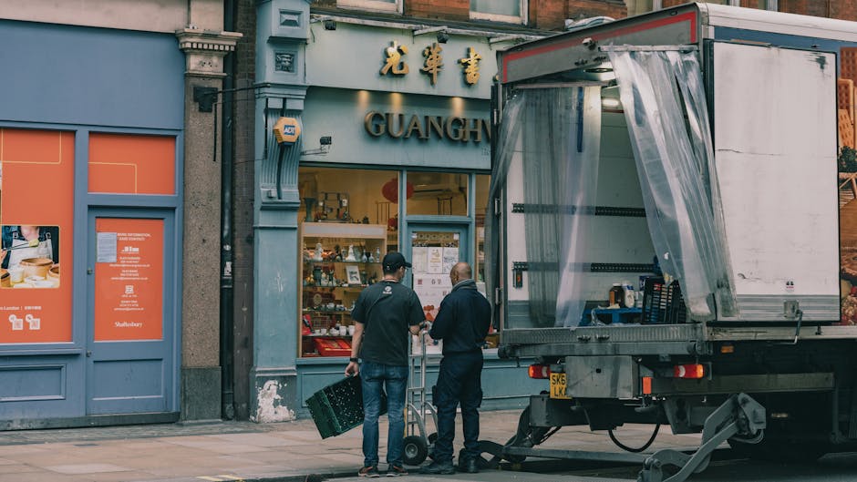 Two workers from Man and Van Chelsea are preparing a home relocation outside a Chinese goods store with the sign 'GUANGH' visible above the entrance. The workers, dressed in dark clothing, are loading packing supplies or small furniture items into a large, partially open, silver moving truck parked on the pavement. Inside the truck, there are visible plastic wrapping materials and stacked black crates, indicative of packing and furniture transport processes. One worker is using a hand trolley to carry a box or item towards the truck, while the other seems to be coordinating or guiding the loading process. The store's window display features various Chinese ceramics and souvenirs arranged on shelves, with some items partially visible through the large glass. The building façade shows typical urban architecture with decorative stonework around the door and window. This scene captures elements of professional removals and packing activities essential to house and furniture removals in Chelsea, reflecting the logistical steps involved in moving services as outlined in the Chelsea removals pricing guide on manandvanchelsea.org.