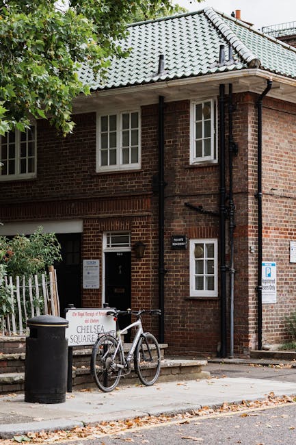 The exterior of a brick residential building with a light grey tiled pitched roof and white-framed windows, situated in Chelsea. In front of the building, a bicycle is parked on the sidewalk near a large black trash bin. To the left, there is a white gate and greenery, including trees partially blocking the view of the building. A sign indicating 'CHESLEA SQUARE' is placed on the sidewalk, next to a small set of steps leading to the entrance. The scene is captured during daylight hours with natural lighting. This setting reflects the typical environment where house removals or furniture transport might take place, consistent with professional removal services like those offered by Man and Van Chelsea, especially in urban residential areas.
