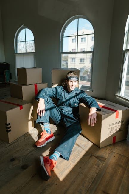 A young man wearing a blue work jumpsuit, orange sneakers, and a headband, sitting on a wooden floor inside a room with large, arched windows allowing natural light to illuminate the space. He is surrounded by several large cardboard boxes sealed with red and black tape, indicating packing for a home relocation. The boxes are arranged in a semi-circle around him, some leaning against the wall and others on the floor, consistent with the packing and moving process carried out by a professional removals service such as Man and Van Chelsea. The room appears spacious with a view of neighboring buildings visible through the windows. This scene captures the loading and packing phase involved in furniture transport and house removals, highlighting the careful handling and organization typical of same day or urgent moves, as often managed by [COMPANY_NAME] in Chelsea.